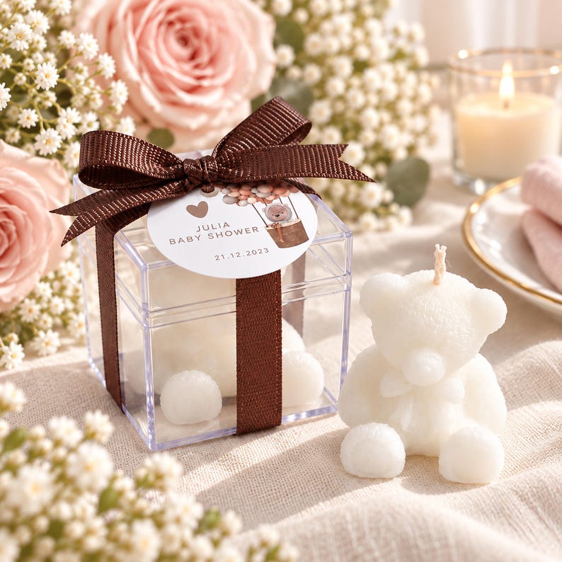 May include: A clear square plastic box containing a white bear-shaped candle, tied with a brown ribbon and a round tag reading Julia Baby Shower 21.12.2023. A matching white bear-shaped candle sits outside the box, featuring a central wick.