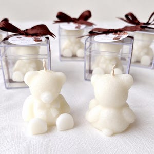 May include: Off-white bear-shaped candles with a single wick. Two candles sit in the foreground, while four additional bear candles are enclosed in clear square plastic boxes, each topped with a brown ribbon and a small circular tag.