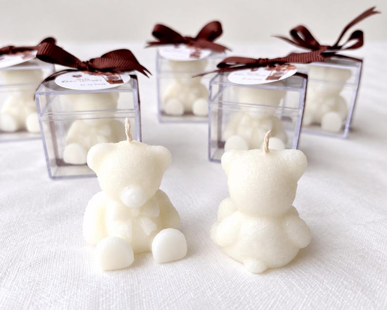 May include: Off-white bear-shaped candles with a single wick. Two candles sit in the foreground, while four additional bear candles are enclosed in clear square plastic boxes, each topped with a brown ribbon and a small circular tag.