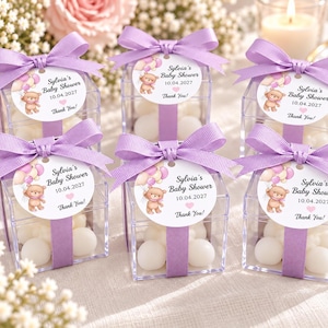 May include: Six clear square plastic favor boxes filled with white candies, each wrapped with a lavender ribbon and a bow. A round tag attached to each box features a cartoon teddy bear with balloons and the text Sylvia's Baby Shower 10.04.2027 Thank You!