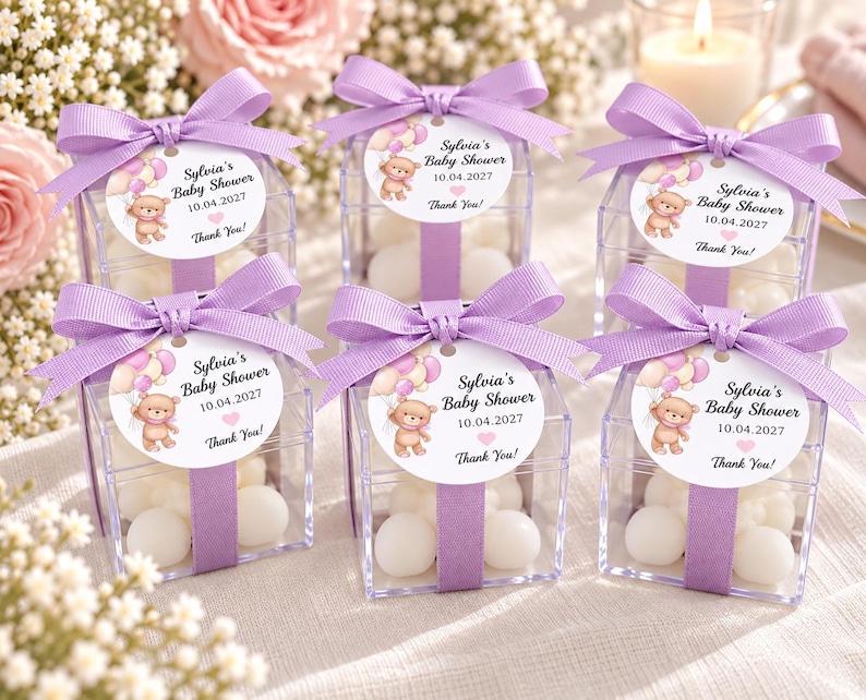 May include: Six clear square plastic favor boxes filled with white candies, each wrapped with a lavender ribbon and a bow. A round tag attached to each box features a cartoon teddy bear with balloons and the text Sylvia's Baby Shower 10.04.2027 Thank You!
