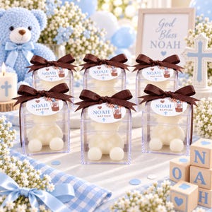 May include: Six clear square boxes each containing a white bear-shaped candle. Each box is topped with a brown ribbon and a tag that reads NOAH BAPTISM with a graphic of a bear in a hot air balloon. The boxes are arranged on a light blue surface.