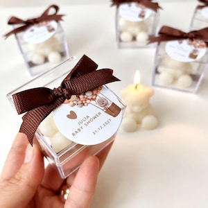 May include: A clear square plastic box containing a white bear-shaped candle, topped with a brown satin ribbon and a circular tag featuring a hot air balloon illustration, the text JULIA BABY SHOWER, and the date 21.12.2027 printed in brown.