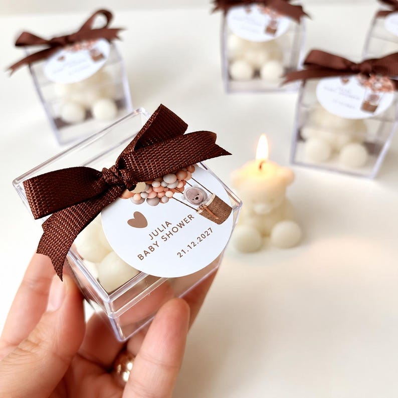 May include: A clear square plastic box containing a white bear-shaped candle, topped with a brown satin ribbon and a circular tag featuring a hot air balloon illustration, the text JULIA BABY SHOWER, and the date 21.12.2027 printed in brown.