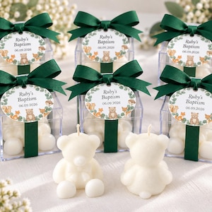 May include: Six clear square favor boxes filled with white candies, each topped with a dark green ribbon bow and an oval tag reading Ruby's Baptism 06.09.2026 with woodland animal illustrations. Two white bear-shaped candles sit in the foreground.