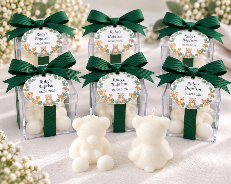 May include: Six clear square favor boxes filled with white candies, each topped with a dark green ribbon bow and an oval tag reading Ruby's Baptism 06.09.2026 with woodland animal illustrations. Two white bear-shaped candles sit in the foreground.