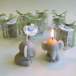 May include: Two gray elephant-shaped candles, one lit with a flame, positioned in front of several clear square gift boxes containing similar elephant candles. Each box is tied with a green ribbon and a round tag reading Chela's Baby Shower.
