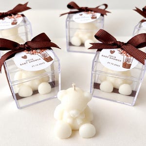 May include: Five clear square boxes containing white teddy bear candles, each topped with a brown ribbon and a circular tag reading JULIA BABY SHOWER 21.12.2023 with a bear in a hot air balloon graphic. A single white teddy bear candle sits in front.