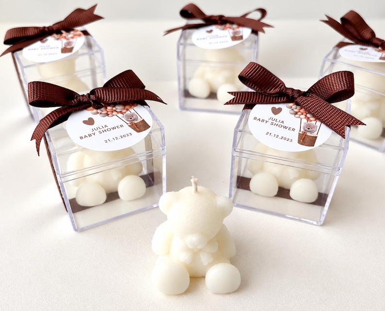 May include: Five clear square boxes containing white teddy bear candles, each topped with a brown ribbon and a circular tag reading JULIA BABY SHOWER 21.12.2023 with a bear in a hot air balloon graphic. A single white teddy bear candle sits in front.