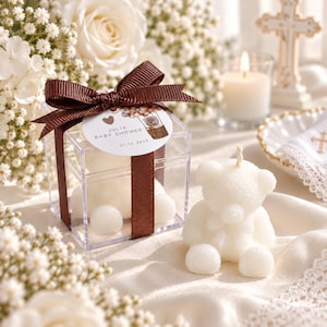 May include: A clear square plastic box containing a white bear-shaped candle, tied with a brown ribbon and a round tag reading Julia Baby Shower 21.12.2023. Beside it sits an identical white bear-shaped candle with a visible wick on top.