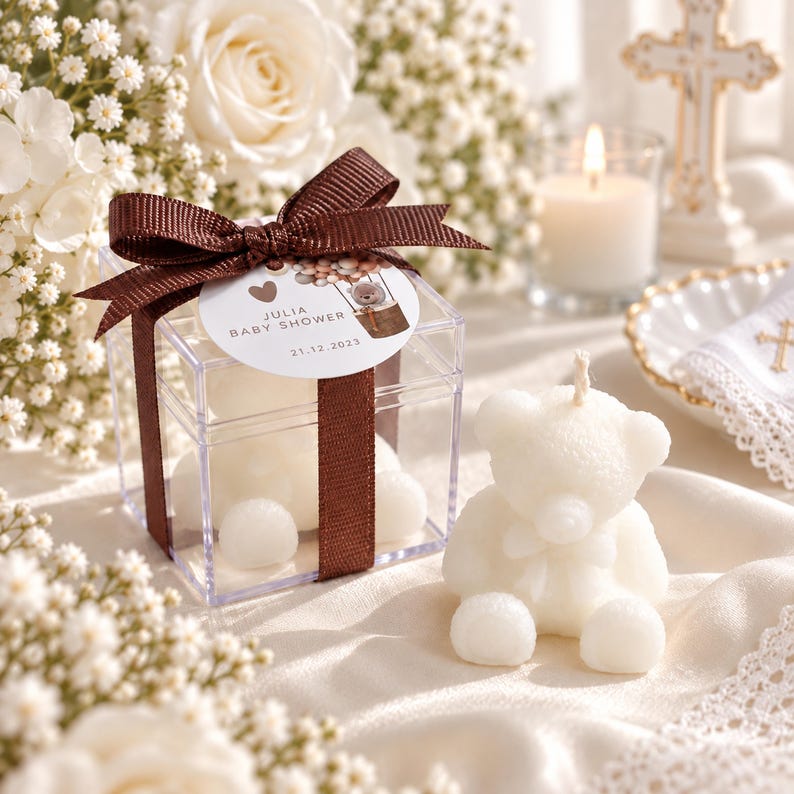 May include: A clear square plastic box containing a white bear-shaped candle, tied with a brown ribbon and a round tag reading Julia Baby Shower 21.12.2023. Beside it sits an identical white bear-shaped candle with a visible wick on top.
