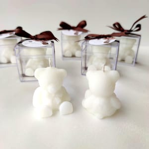 May include: A collection of white bear-shaped candles, some placed outside clear square boxes and others inside, each box topped with a brown ribbon and a small circular tag. The candles feature a seated bear design with a visible wick on top.
