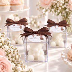 May include: Three clear square plastic favor boxes each containing a white teddy bear-shaped candle. A brown satin ribbon bow is tied on top of each box with a round white tag printed with a heart, balloons, and the text Julia Baby Shower.