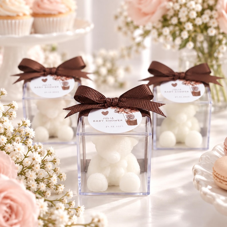 May include: Three clear square plastic favor boxes each containing a white teddy bear-shaped candle. A brown satin ribbon bow is tied on top of each box with a round white tag printed with a heart, balloons, and the text Julia Baby Shower.