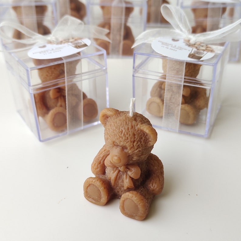 以下が含まれることがあります： 透明なプラスチック製の箱に入った、小さな茶色のテディベアの形をしたろうそく。ろうそくは白い表面の上に置かれています。箱には白いリボンと小さなタグが付いており、テキストが書かれています。