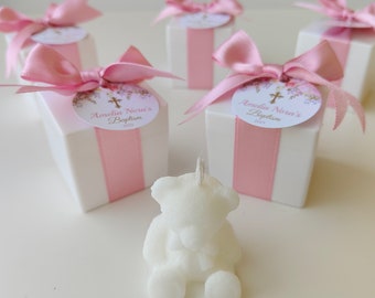 May include: A white bear-shaped candle sits in front of several white square gift boxes wrapped with pink satin ribbons and bows. Each box features a round tag with floral illustrations, a gold cross, and the text Amelia Nora's Baptism 2025.
