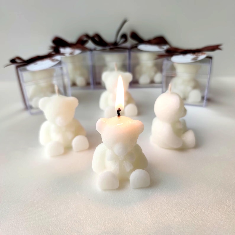 May include: A collection of white bear-shaped candles, with one lit in the center. Several candles are enclosed in clear square boxes topped with brown ribbon bows. The candles are molded into the shape of seated teddy bears with smooth surfaces.