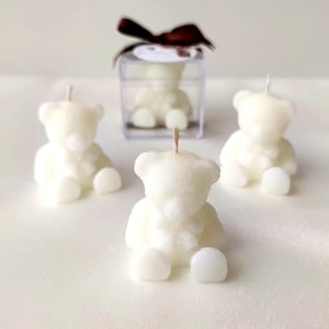 May include: Three off-white bear-shaped candles with a single wick each, positioned in a triangular arrangement. A fourth identical bear candle is enclosed within a clear square plastic box topped with a dark brown ribbon bow.