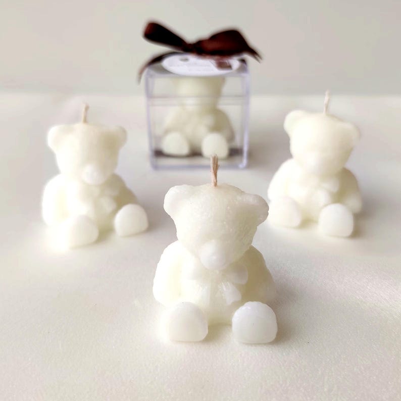 May include: Three off-white bear-shaped candles with a single wick each, positioned in a triangular arrangement. A fourth identical bear candle is enclosed within a clear square plastic box topped with a dark brown ribbon bow.