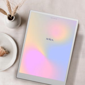 May include: A digital tablet displays a soft, colorful gradient with the word "notes." A small white vase holds wheat stalks, and a plate with a piece of cake sits nearby. The tablet has a silver frame.