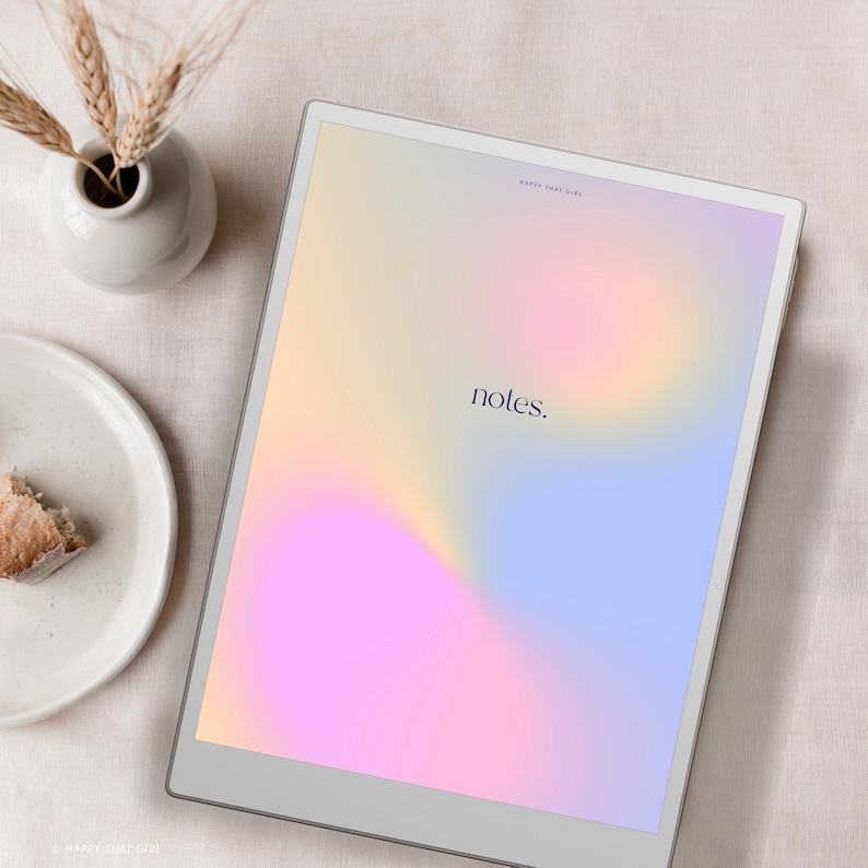 May include: A digital tablet displays a soft, colorful gradient with the word "notes." A small white vase holds wheat stalks, and a plate with a piece of cake sits nearby. The tablet has a silver frame.