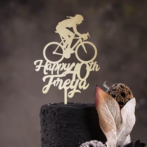 May include: Gold-colored wooden cake topper featuring a silhouette of a person riding a bicycle with the text "Happy 40th Freya" below.