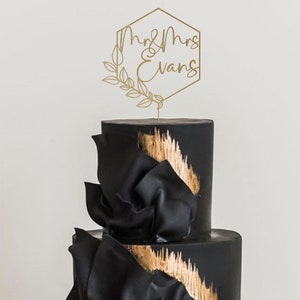 May include: Gold geometric cake topper with the text "Mr & Mrs Evans" and a leafy design. The topper is on top of a black and gold wedding cake.
