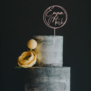 May include: A two-tiered grey wedding cake with yellow flowers and a rose gold cake topper that says "Emma & Mark" in cursive.