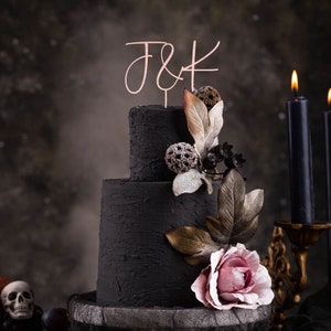 Peut inclure: Un gâteau de mariage noir à deux étages décoré d'une rose rose et d'accents en feuilles d'or. Le gâteau est surmonté d'un topper en or rose qui dit "J&K".