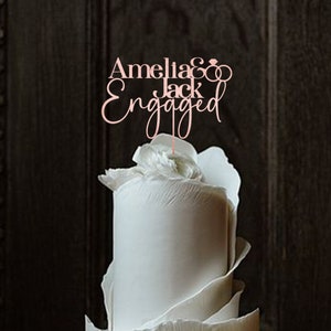 May include: A white wedding cake with a topper that says "Amelia & Jack Engaged" with two intertwined rings.
