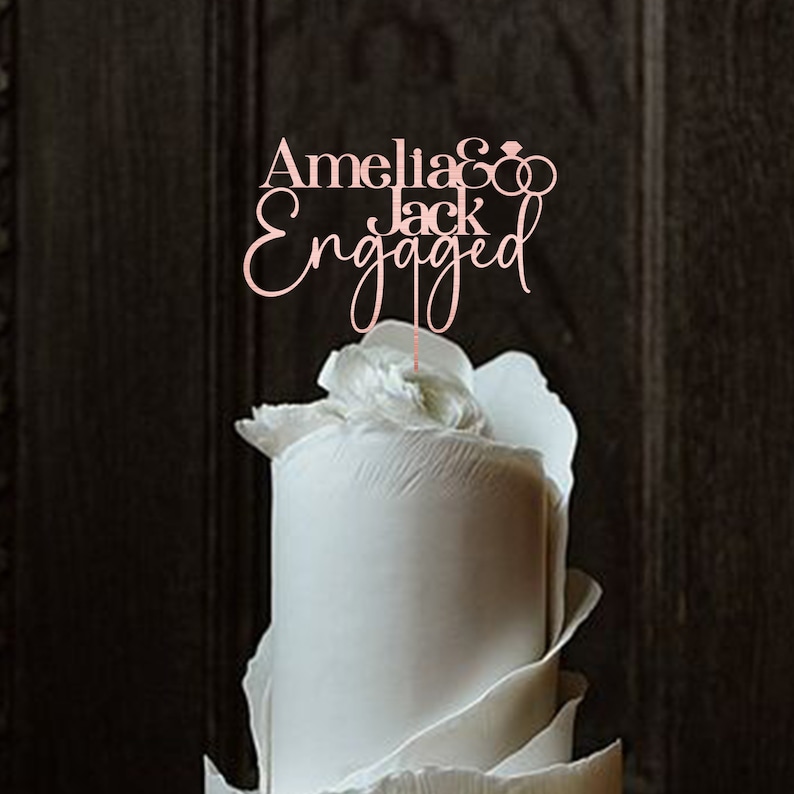 May include: A white wedding cake with a topper that says "Amelia & Jack Engaged" with two intertwined rings.