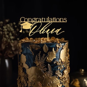 Op de afbeelding: Een goudkleurige taarttopper met de tekst Congratulations en de naam Olivia in een sierlijk lettertype, vergezeld van een icoon van een afstudeerhoed. De topper staat boven op een donkerblauwe taart versierd met bladgoud en bloemaccenten.