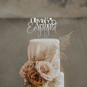 May include: A white wedding cake with a silver topper that says "Olivia & Arthur Engaged" with two interlocking rings. The cake is decorated with brown and pink roses.