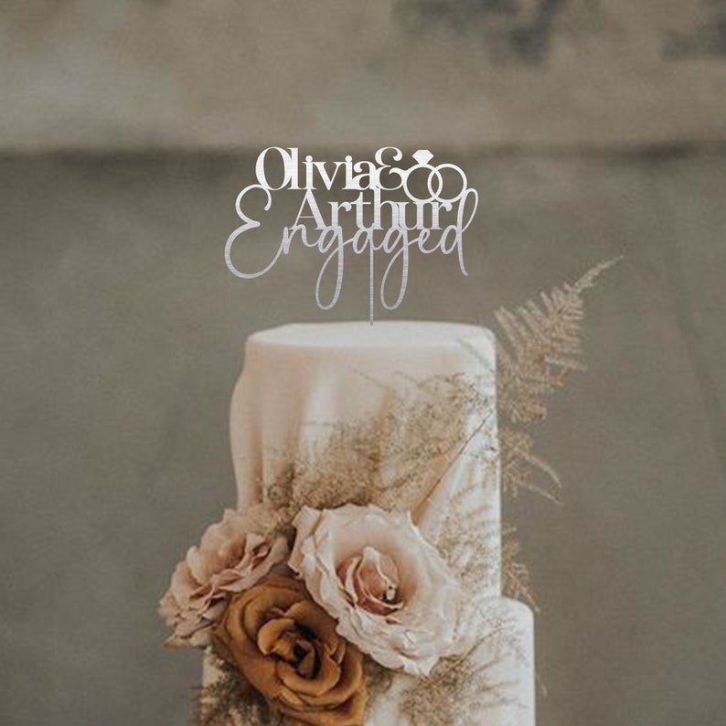 May include: A white wedding cake with a silver topper that says "Olivia & Arthur Engaged" with two interlocking rings. The cake is decorated with brown and pink roses.