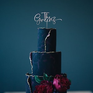 May include: A four-tiered dark blue wedding cake with gold accents and red and pink flowers. The cake topper says "The Greens" in a cursive font.