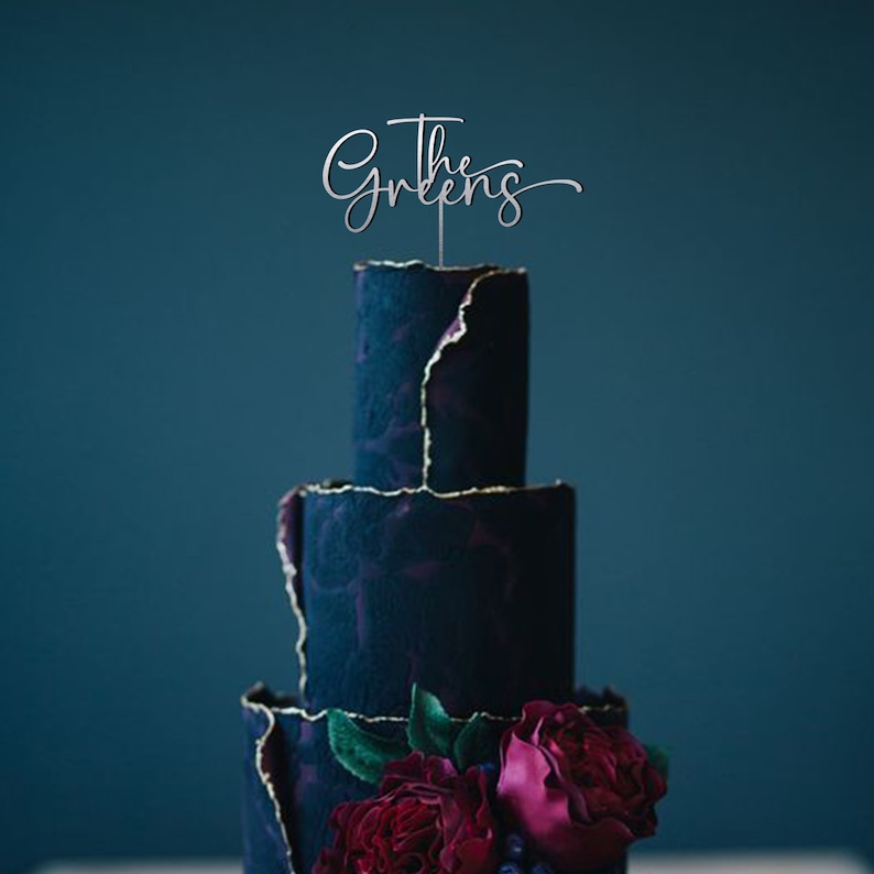 May include: A four-tiered dark blue wedding cake with gold accents and red and pink flowers. The cake topper says "The Greens" in a cursive font.