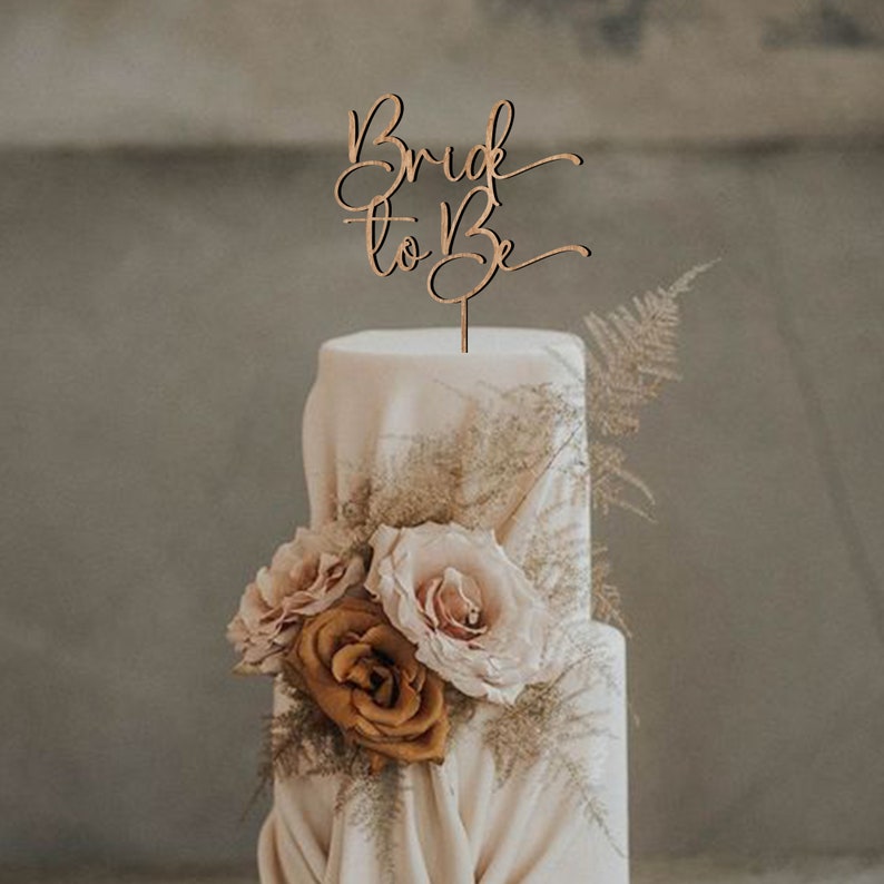 May include: A white wedding cake with a wooden cake topper that says "Bride to Be" in cursive. The cake is decorated with brown and pink roses and greenery.