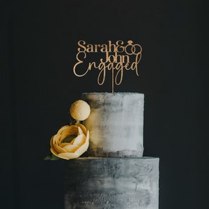 May include: A wooden cake topper with the text "Sarah & John Engaged" and two intertwined rings. The topper is on a two-tiered gray cake with yellow flowers.