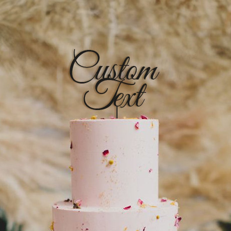 May include: A two-tiered pink cake decorated with dried flowers and a black cake topper. The cake topper reads "Custom Text" in an elegant script font. The cake is set against a blurred, neutral background.
