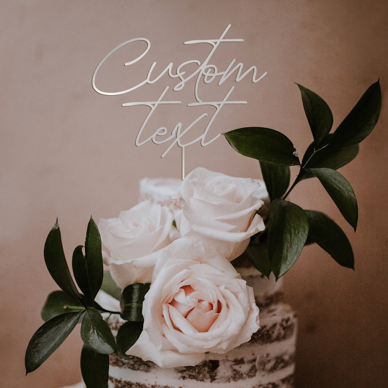 May include: A cake topper with the words "Custom Text" in a cursive font. The topper is silver and is placed on top of a cake decorated with white roses and green leaves. The cake has a light brown, textured exterior.