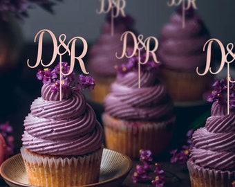Pode incluir: Cupcakes roxos com cobertura branca cobertos com toppers de cupcake em ouro rosa que dizem "D&R".