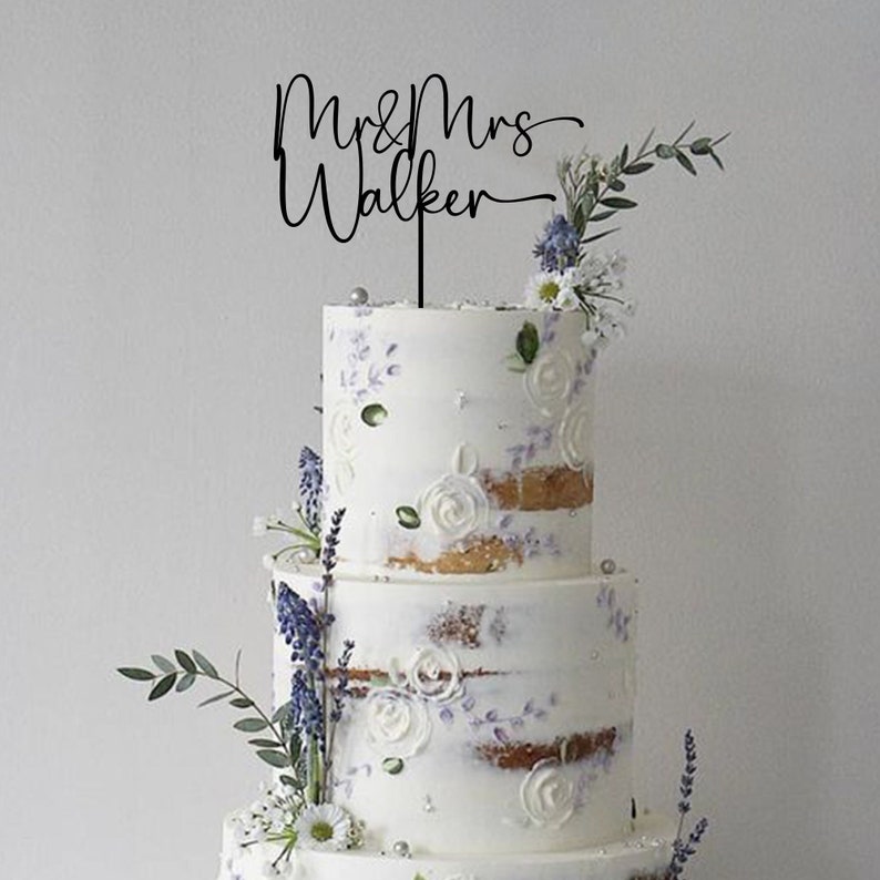 May include: A white wedding cake with a black acrylic cake topper that says "Mr & Mrs Walker" in cursive. The cake is decorated with white frosting and purple flowers.