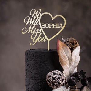 Puede incluir: Topper de pastel dorado con el texto "We Will Miss You" y un corazón con el nombre "Sophia" en su interior. El topper está sobre un pastel negro.