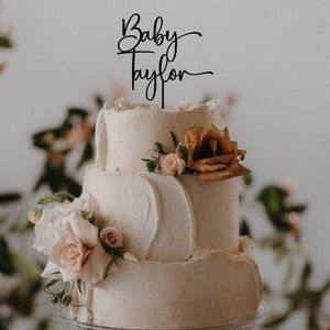 May include: A three-tiered wedding cake decorated with buttercream frosting and fresh flowers. The cake topper says "Baby Taylor" in a cursive font.