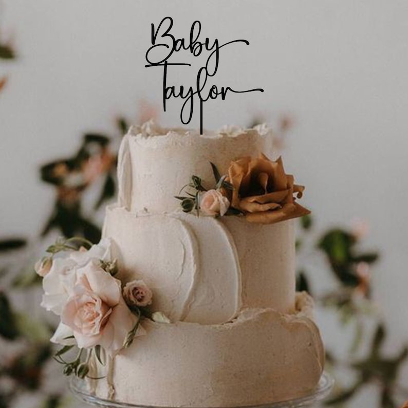 May include: A three-tiered wedding cake decorated with buttercream frosting and fresh flowers. The cake topper says "Baby Taylor" in a cursive font.