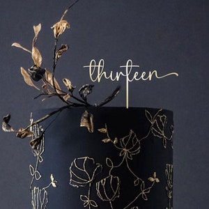 May include: Gold cake topper with the word "thirteen" in a cursive font. The topper is on top of a black cake with gold floral designs.