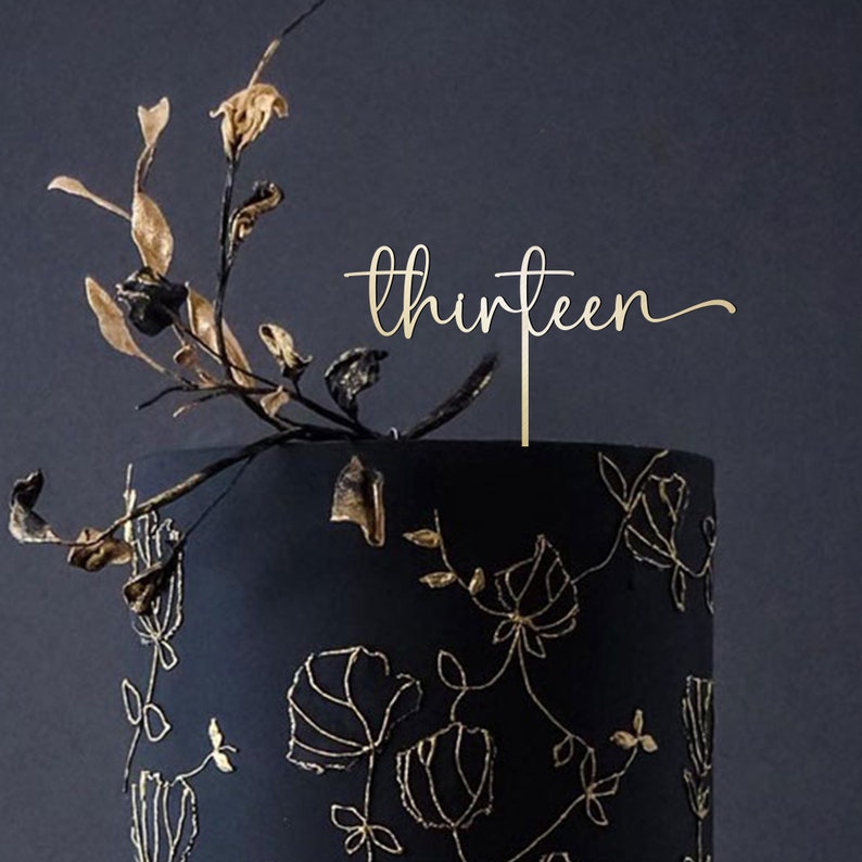 May include: Gold cake topper with the word "thirteen" in a cursive font. The topper is on top of a black cake with gold floral designs.