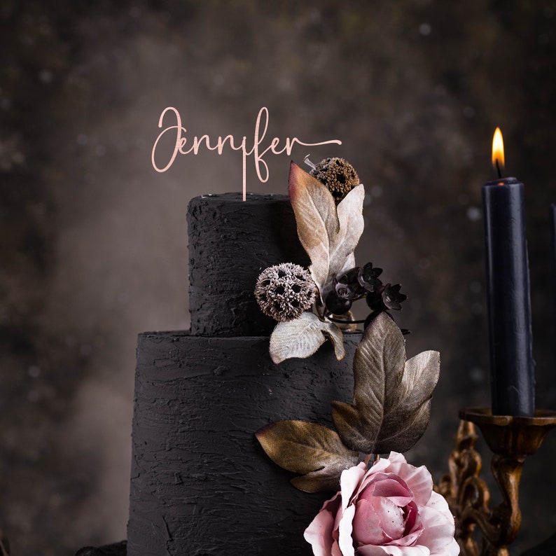 May include: A two-tier black wedding cake decorated with dried flowers and greenery. The cake is topped with a pink and gold topper that says "Jennifer".