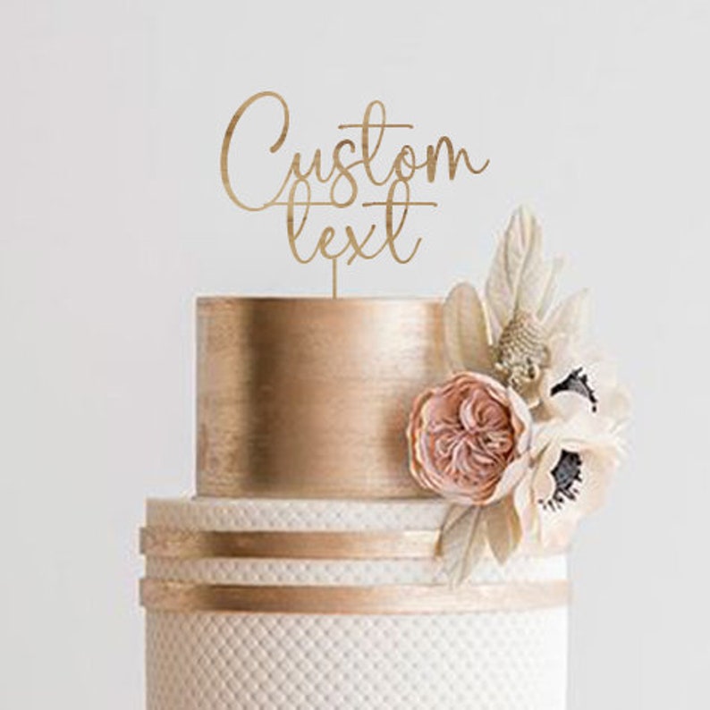 May include: A two-tiered wedding cake with gold accents and floral decorations. The cake features a topper with the words "Custom Text" in a delicate script. The cake is decorated with a textured white base and gold ribbons.