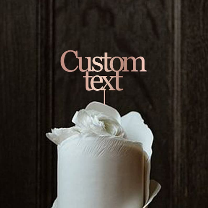 May include: A rose gold cake topper with the words "Custom Text" in a stylish font. The topper is placed on top of a white frosted cake with a white flower decoration. The background is a dark wood.